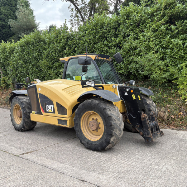 Yellow CAT skid-steer loader with a front bucket parked on a paved lot, dense green hedge in the background.
