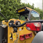 Rear view of a yellow construction machine with exposed hydraulic hoses, warning stickers, and a red taillight set against green hedges.