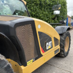 Close-up of a yellow CAT construction vehicle, showing the front panel, large tire, and side mirror with a hedge and houses in the background.