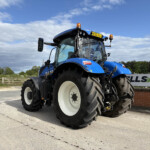 Blue agricultural tractor seen in a parking area, front-left angle with oversized tires and orange beacon lights on top panel license plate AE20 AJJ