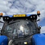 Front view of a blue tractor cab with orange beacon lights and license plate AE20 AUJ against a cloudy sky.