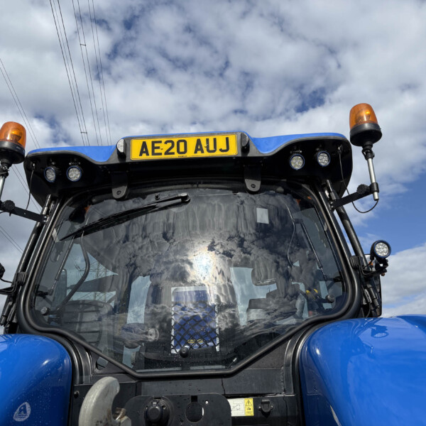 Front view of a blue tractor cab with orange beacon lights and license plate AE20 AUJ against a cloudy sky.
