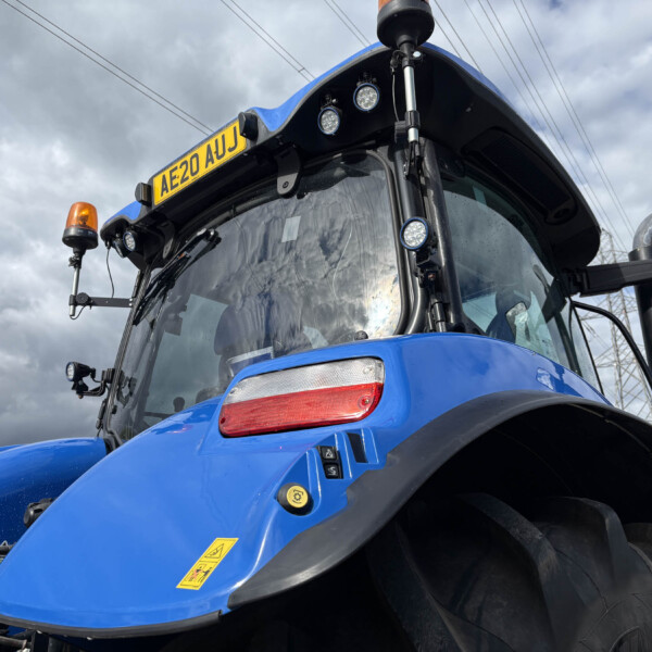 Front close-up of a blue tractor cab with an orange beacon, license plate AE20 AUJ, and multiple exterior lights against a cloudy sky.
