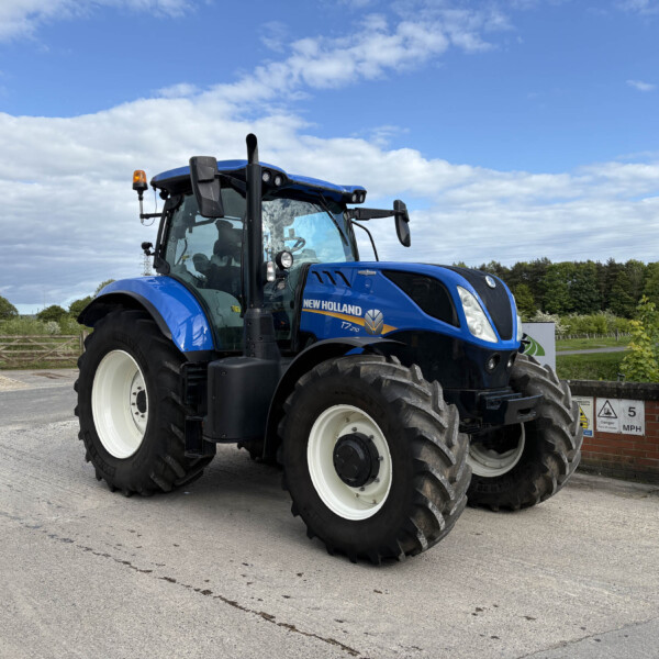 Blue New Holland tractor parked in an open lot with a green fence and cloudy blue sky.