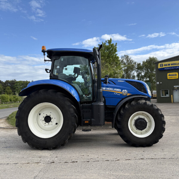 Front-left close-up of a blue New Holland tractor with large white-walled tires, parked beside a New Holland Agriculture building.