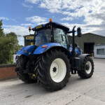 Blue New Holland tractor with oversized white wheels parked outdoors beside a warehouse under a partly cloudy sky.