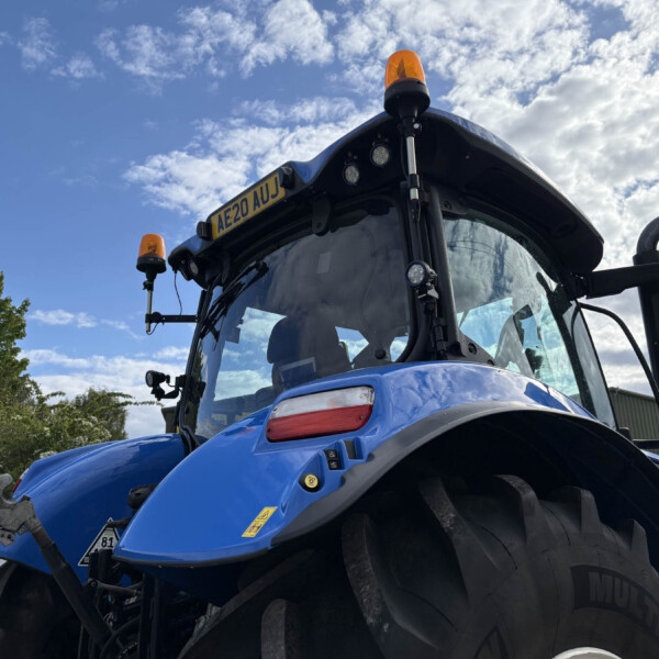Front view of a blue agricultural tractor with orange warning beacon on top and license plate AE20 AUJ against a blue sky.