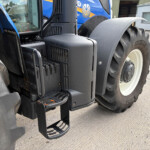 Close-up of a large tractor showing black metal steps, wheel well, and blue cab with thick treaded tire nearby on a concrete surface.