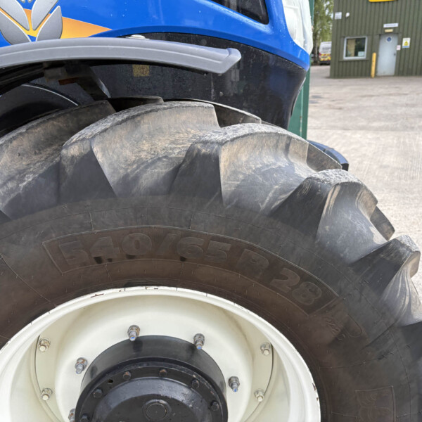 Close-up of a large tire on a blue tractor, showing muddy tread and part of the vehicle body with a yellow–gray logo in an outdoor industrial setting.
