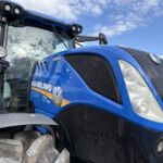 Close-up of a blue New Holland tractor front, showing the grille, large front tire treads, and part of the cab with lights and mirrors under a blue sky.