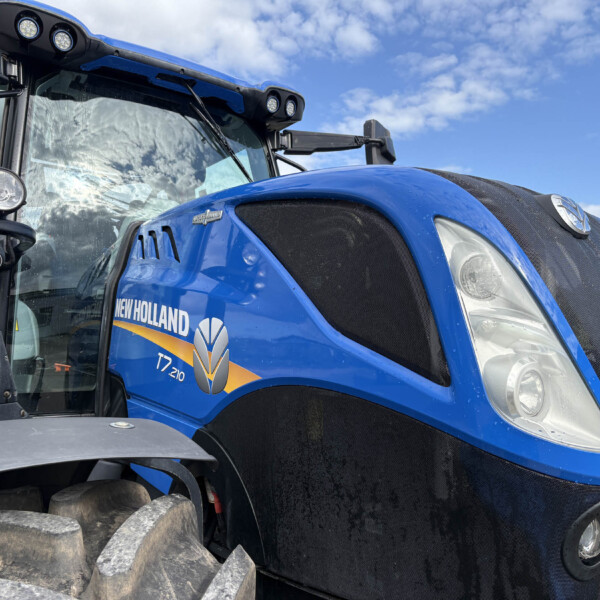 Close-up of a blue New Holland tractor front, showing the grille, large front tire treads, and part of the cab with lights and mirrors under a blue sky.