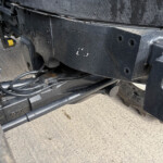 Close-up of a dirty black vehicle frame with hoses and wiring beneath the undercarriage of a truck/piece of heavy machinery.