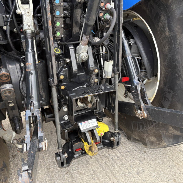 Close-up view of a tractor hitch and hydraulic assembly with hoses, metal arms, and a yellow towing hook near a large tire; utility controls and cables are visible in a dirty workshop area.