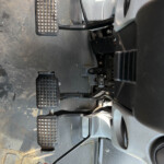 Inside a dusty vehicle cab showing three pedals (clutch, brake, accelerator) with a red pedal pad nearby and part of the steering wheel visible.