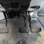 Inside a vehicle's driver footwell showing three pedals with textured rubber pads and an orange-accent pedal, with wiring and metal linkage visible.