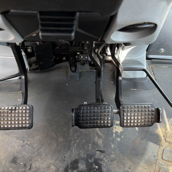 Inside a vehicle's driver footwell showing three pedals with textured rubber pads and an orange-accent pedal, with wiring and metal linkage visible.