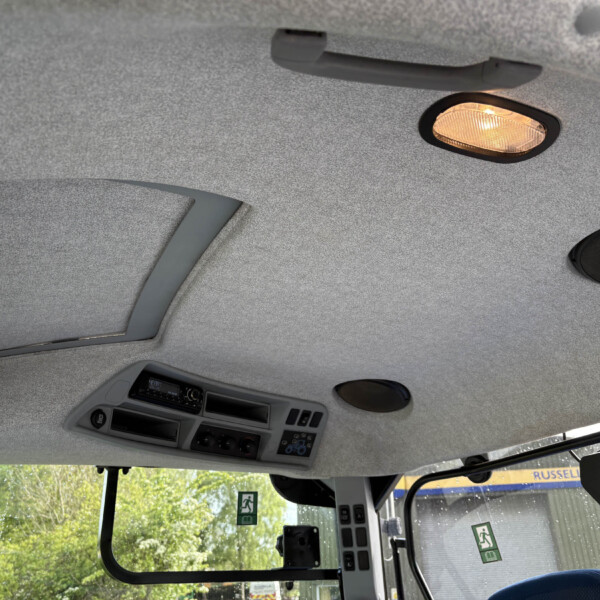Interior of a vehicle cab ceiling with a lit overhead light, sun visor, and control panel; emergency exit signs visible outside the window.
