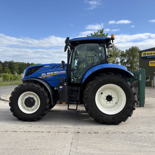 Blue New Holland T7 tractor parked beside a New Holland Agriculture service building, large tires and clear blue sky above