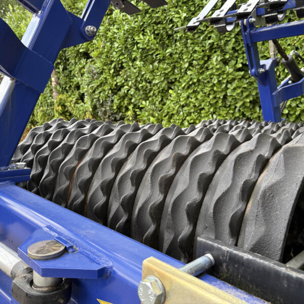 Close-up of a blue metal framework with large black roller rollers, outdoors over a ribbed roof and green hedge in the background