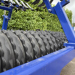 Close-up of blue metal frame and large ribbed tires on a trailer, with hedge and a brick house in the background outside.