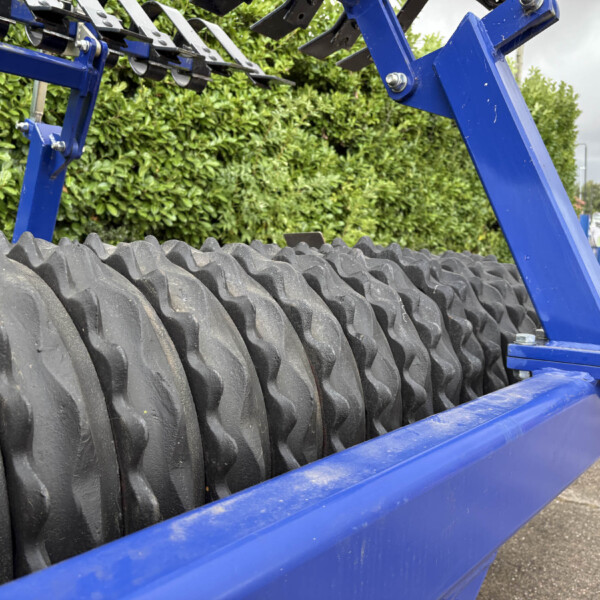 Close-up of blue metal frame and large ribbed tires on a trailer, with hedge and a brick house in the background outside.