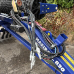 Close-up of hydraulic hoses and metal fittings on a blue farming implement, with a pressure gauge and yellow warning labels, outdoors by plants.