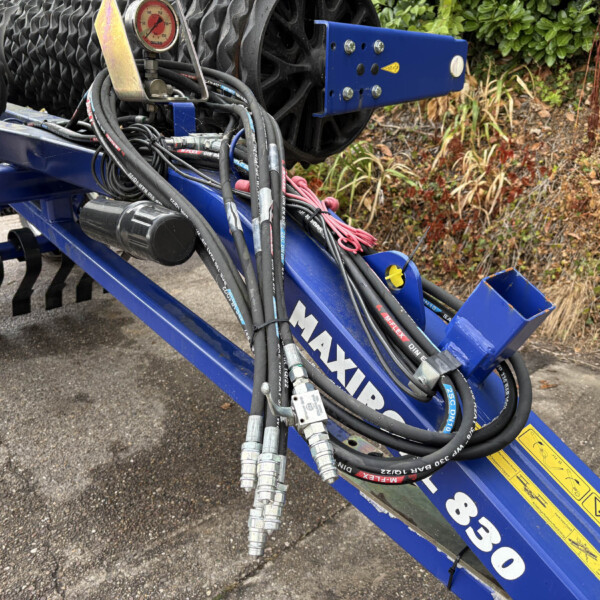 Close-up of hydraulic hoses and metal fittings on a blue farming implement, with a pressure gauge and yellow warning labels, outdoors by plants.