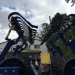 Blue agricultural implement with curved tines, set outdoors in front of a building and hedge.