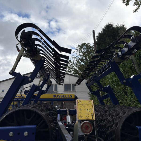Blue agricultural implement with curved tines, set outdoors in front of a building and hedge.