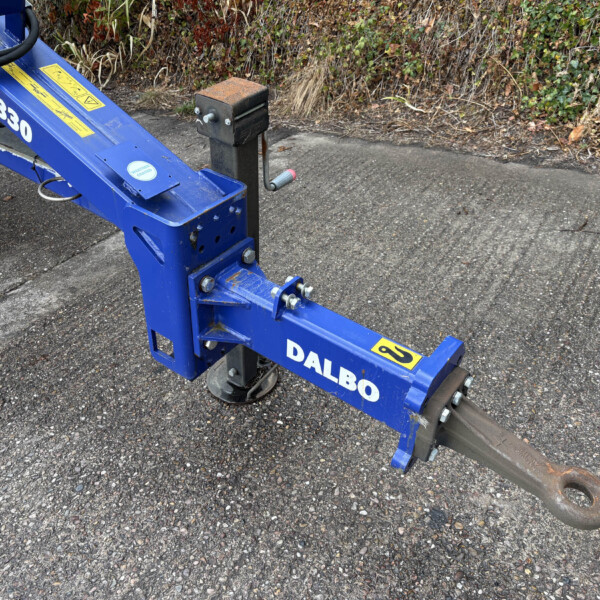 Close-up of a blue hydraulic trailer arm with a curved hitch on a paved surface, ivy behind it.