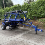 Blue Dalbo cultivator attachment with rolling tines parked in a lot beside a tall hedge, ready for field work.