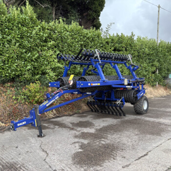 Blue Dalbo seed drill on a trailer parked beside a tall hedge along the street, under a cloudy sky.
