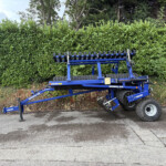 Blue DALBO agricultural implement attached to a trailer parked beside a tall green hedge.
