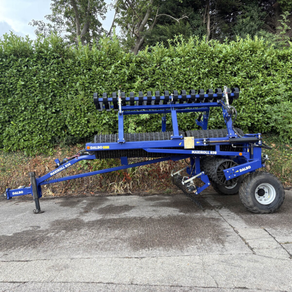 Blue DALBO agricultural implement attached to a trailer parked beside a tall green hedge.