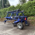 Blue MAXIROLL 830 agricultural roller implement on a trailer, parked beside a dense hedge in a paved lot