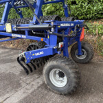 Blue MAXIROLL 830 agricultural implement with roller drums and hydraulic hoses, parked beside a hedge outdoors.