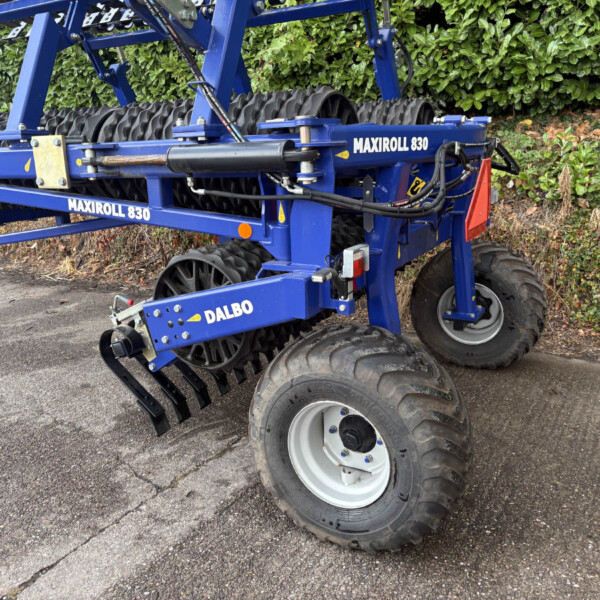Blue MAXIROLL 830 agricultural implement with roller drums and hydraulic hoses, parked beside a hedge outdoors.