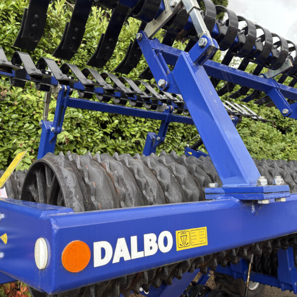 Blue Dalbo agricultural roller/harrow with stacked black circular rollers on a blue steel frame, outdoors beside a hedge.