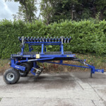 Blue agricultural implement with multiple roller discs parked in front of a tall hedge, overcast sky in background.