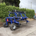 Blue Dalbo MaxiRoll 830 roller harrow attachment on a trailer, parked beside a tall green hedge and an urban street.