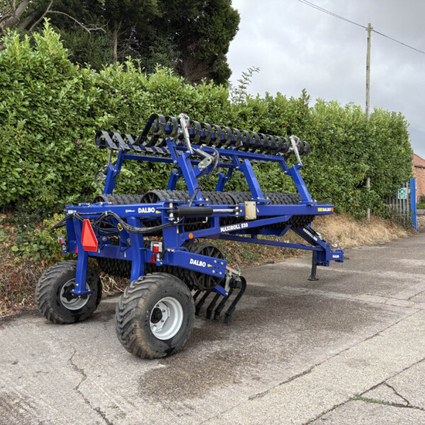 Blue Dalbo MaxiRoll 830 roller harrow attachment on a trailer, parked beside a tall green hedge and an urban street.