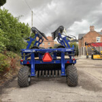 Front view of a large blue Dalbo Maxiroll 830 farming implement with folded tines, parked near a hedge on a cloudy day.