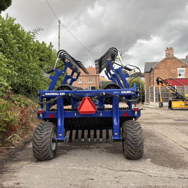 Front view of a large blue Dalbo Maxiroll 830 farming implement with folded tines, parked near a hedge on a cloudy day.