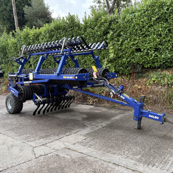 Blue Dalbo cultivator attachment with rolling tines parked in a lot beside a tall hedge, ready for field work.