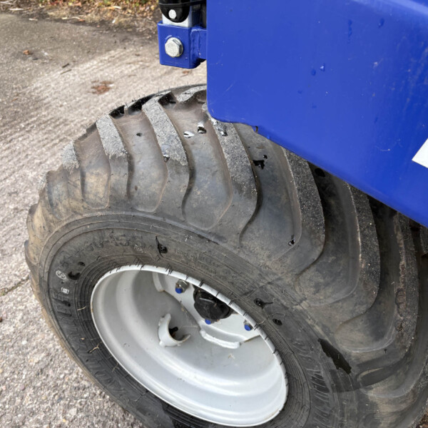 Close-up of a large heavy-machinery wheel: thick treaded tire next to a blue metal frame with a white letter on it, on a paved surface.