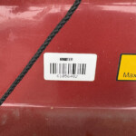 Red metallic surface with a black braided rope crossing diagonally; white barcode label and a partial yellow warning sticker visible.