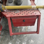 Close-up of a yellow hydraulic cylinder on a red agricultural machine frame, with chains draped over a perforated red work surface.
