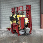 Red agricultural sprayer with yellow tanks parked in front of a gray corrugated metal door outside a warehouse.