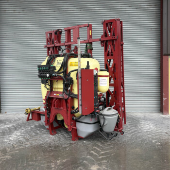 Red agricultural sprayer with yellow tanks parked in front of a gray corrugated metal door outside a warehouse.