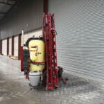 Red agricultural sprayer with a yellow tank standing inside a warehouse with corrugated metal shutters on the right side.